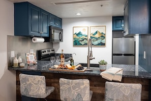 Una cocina con gabinetes azul oscuro, electrodomésticos de acero inoxidable, encimera de mármol y dos cuadros con paisajes.