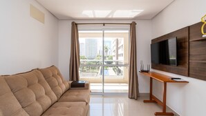 A modern living room with a sofa, a flat-screen TV mounted on a wooden panel, and a sliding glass door leading to a balcony with a city view. - Modernos Flats no Setor Bueno TXP03 (Goiânia)