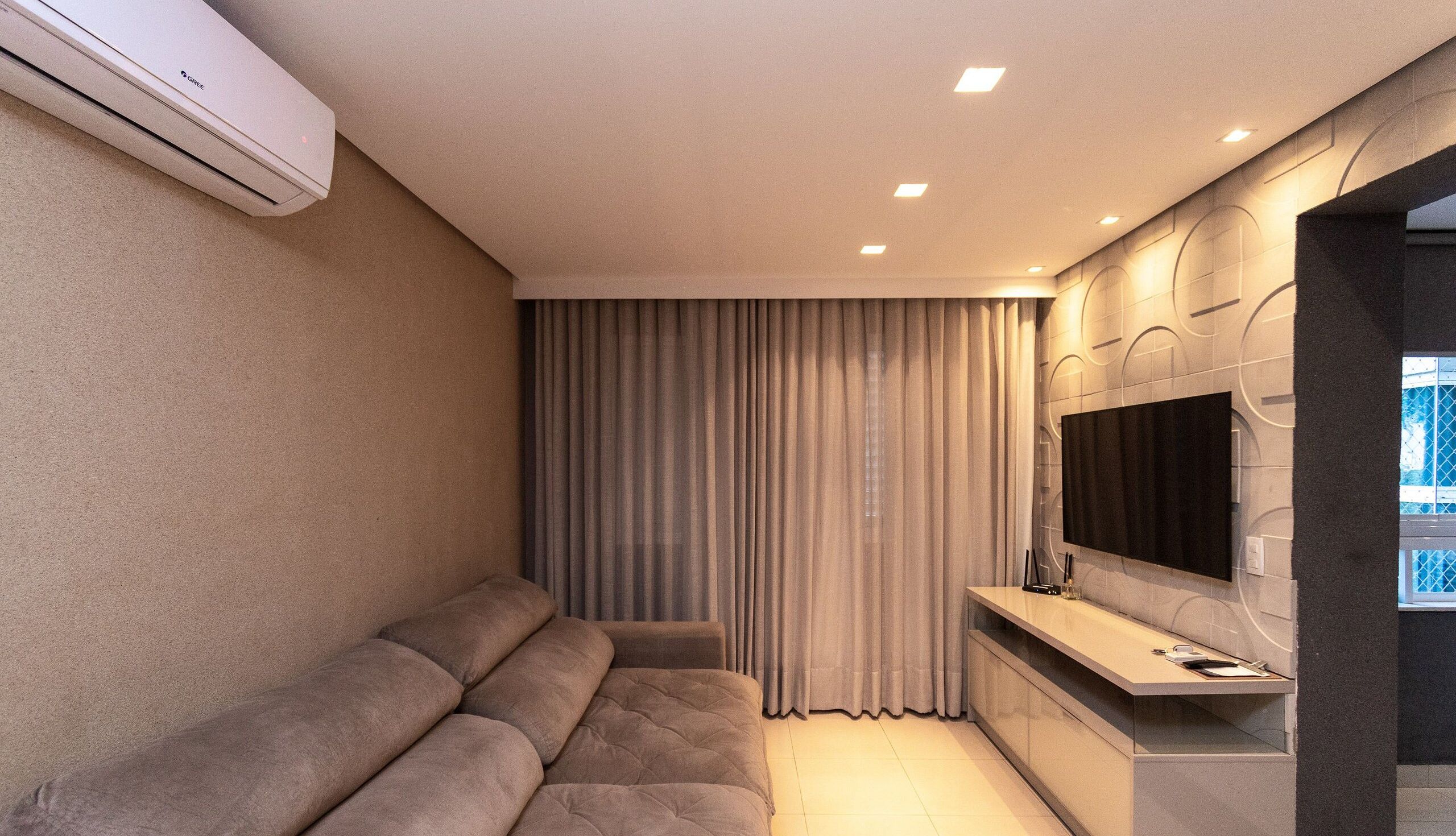 A modern living room with a sofa, a flat-screen TV mounted on the wall, and an air conditioning unit.