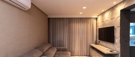 A modern living room with a sofa, a flat-screen TV mounted on the wall, and an air conditioning unit.