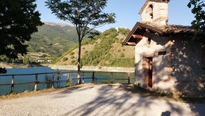 Edifici de pedra rústic amb un petit campanar, un camí de grava que condueix a una tanca de fusta amb vista a un llac, i un paisatge de muntanyes al fons.