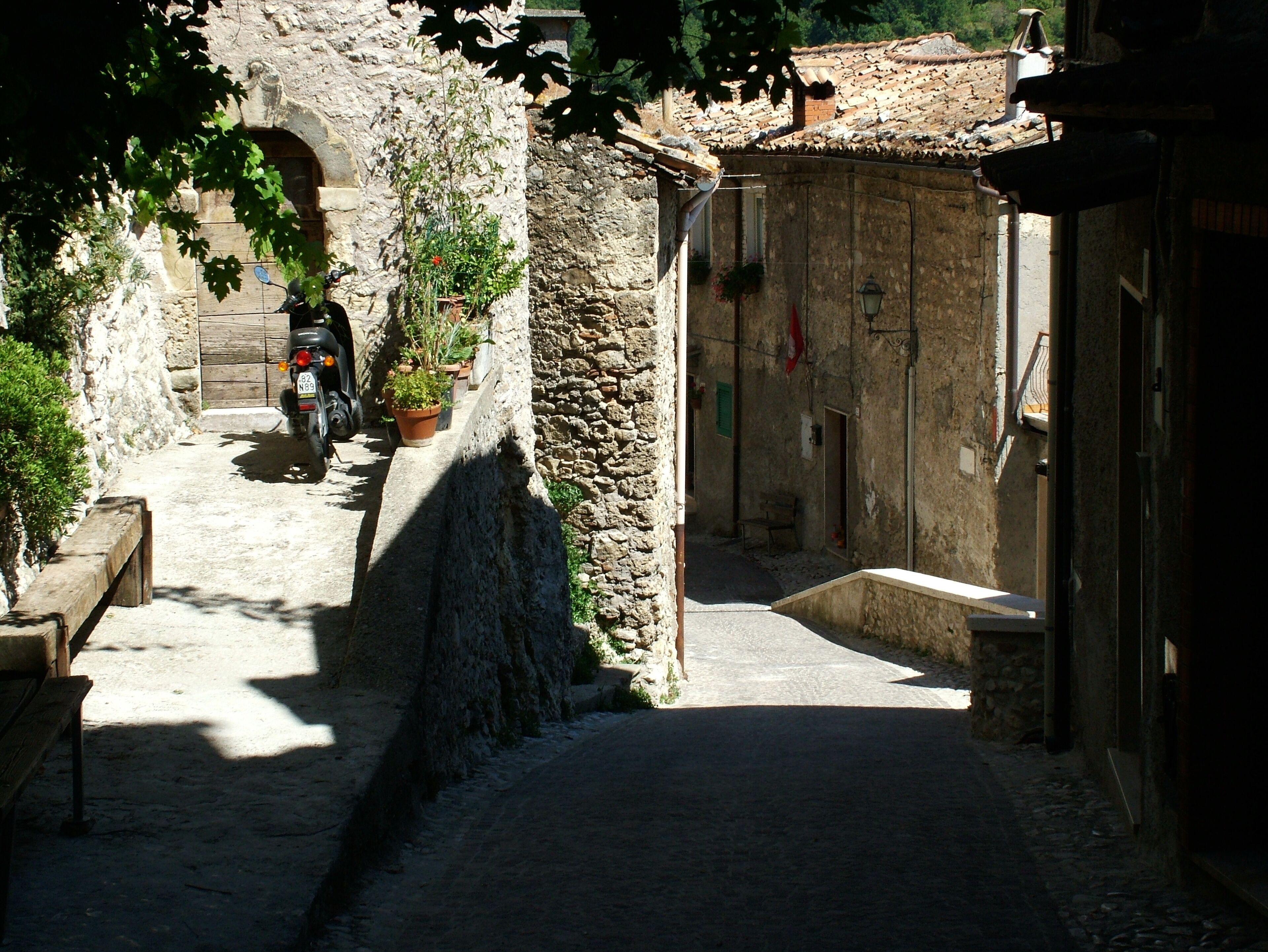 A narrow cobblestone street with a stone wall on one side and a building entrance on the other. A motorcycle is parked near the wall, and there is a bench on the left.