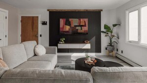 Un salon moderne avec un canapé gris, un pouf noir, un buffet en bois, une grande peinture abstraite et une plante en pot.