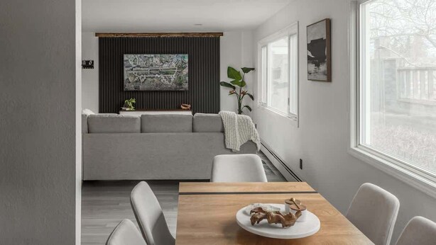 Un salon moderne avec un canapé gris, une table à manger en bois surmontée d’une assiette blanche et une grande fenêtre donnant sur un paysage arboré.