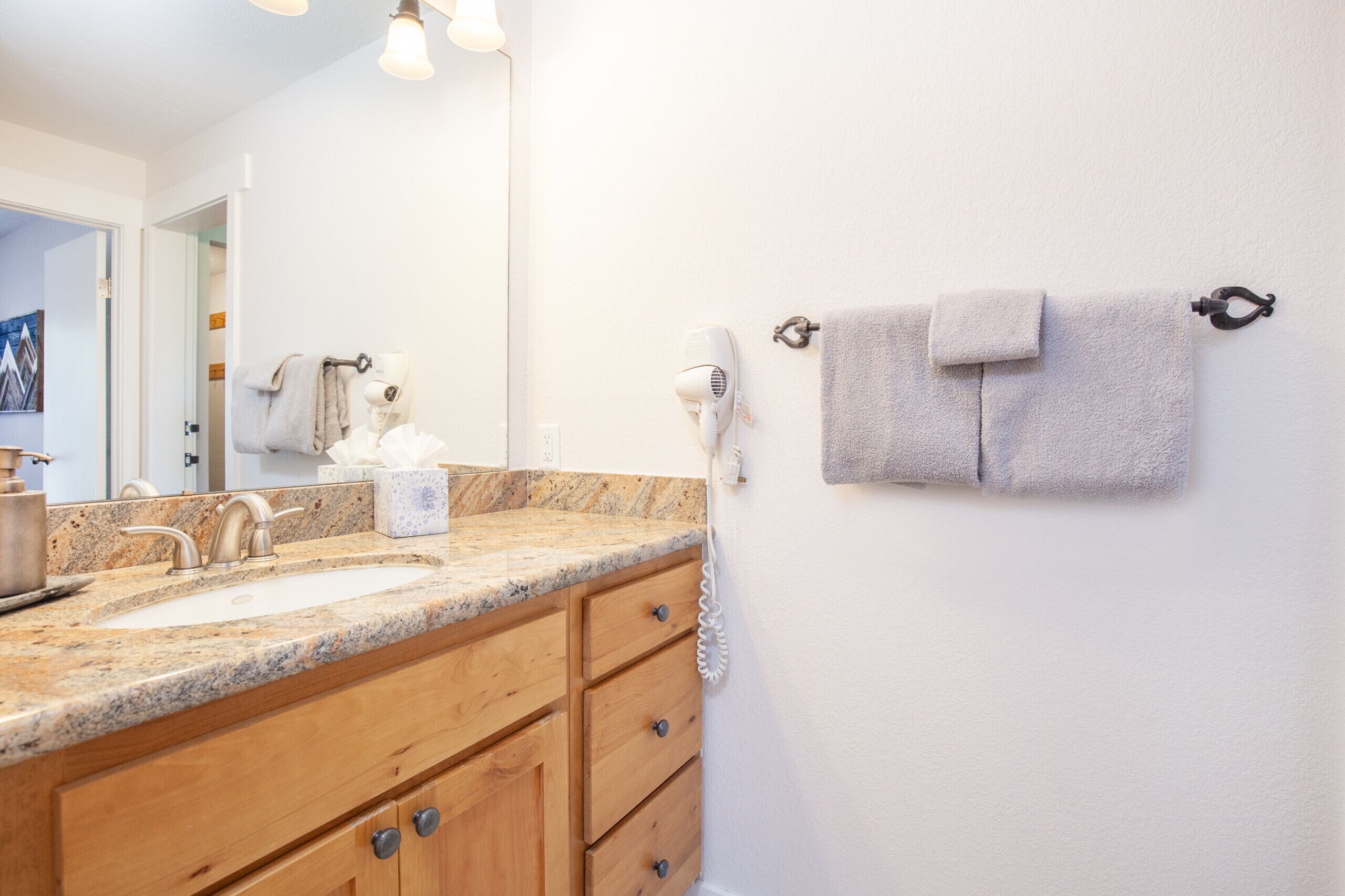 A bathroom with a granite countertop sink, wooden cabinets, a towel rack with two towels, and a hairdryer.