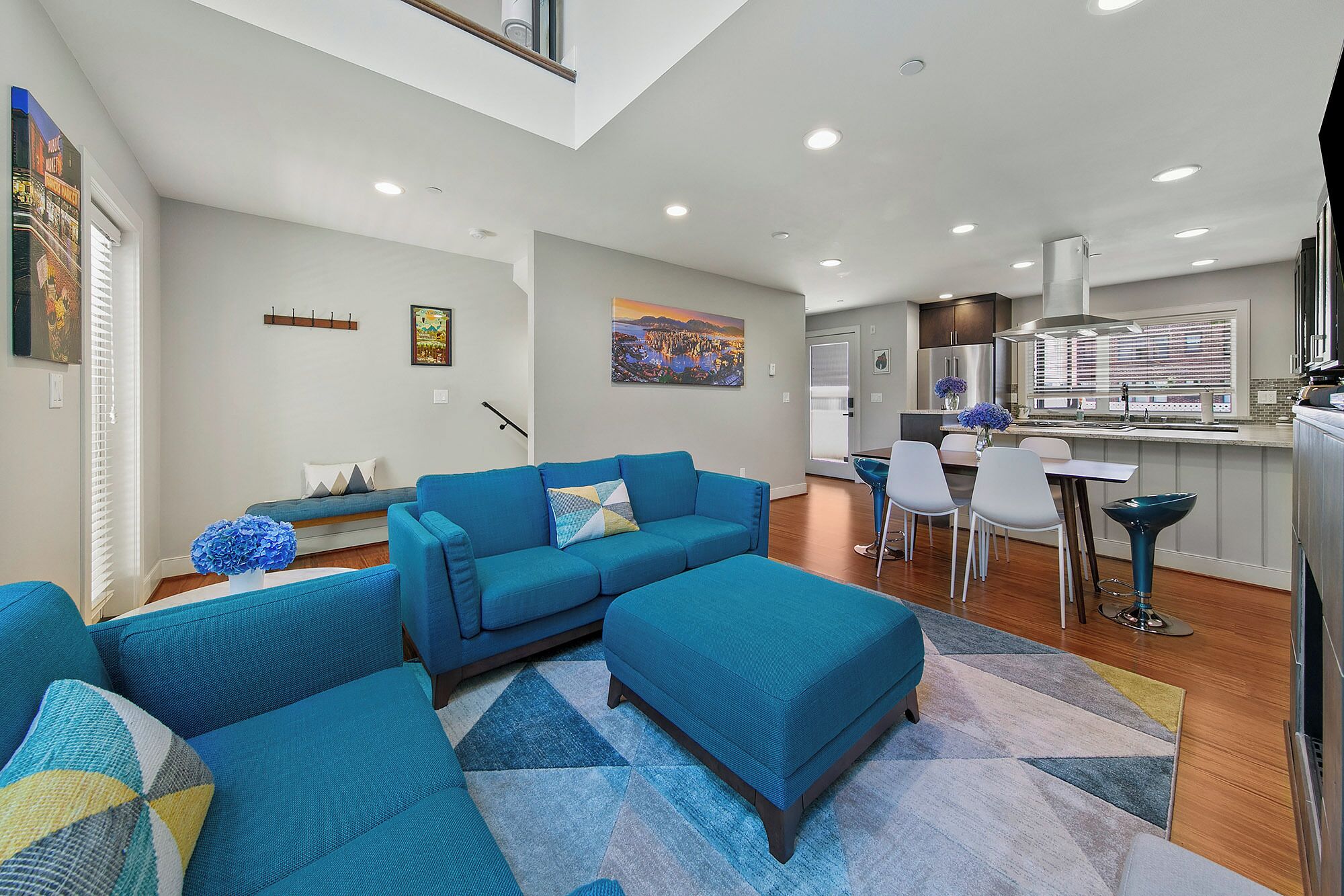 A modern living room with a blue sofa set, a dining area with white chairs, and a kitchen with stainless steel appliances.