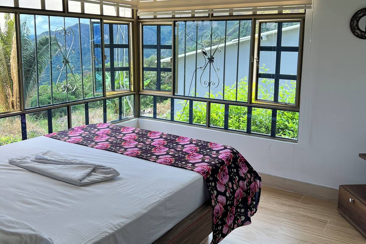 A bedroom with a bed, large windows, and a view of greenery.