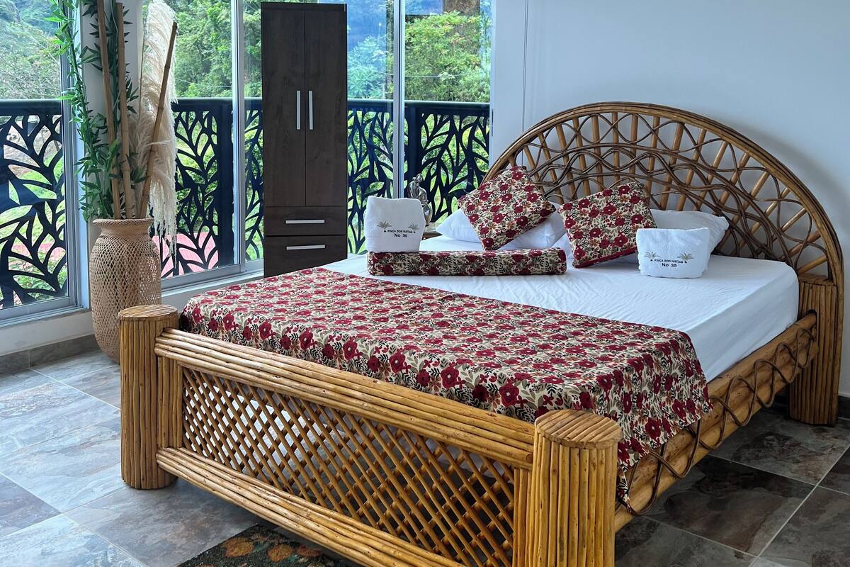 A spacious bedroom with a large bed, a ceiling fan, and a view of lush greenery through large windows.