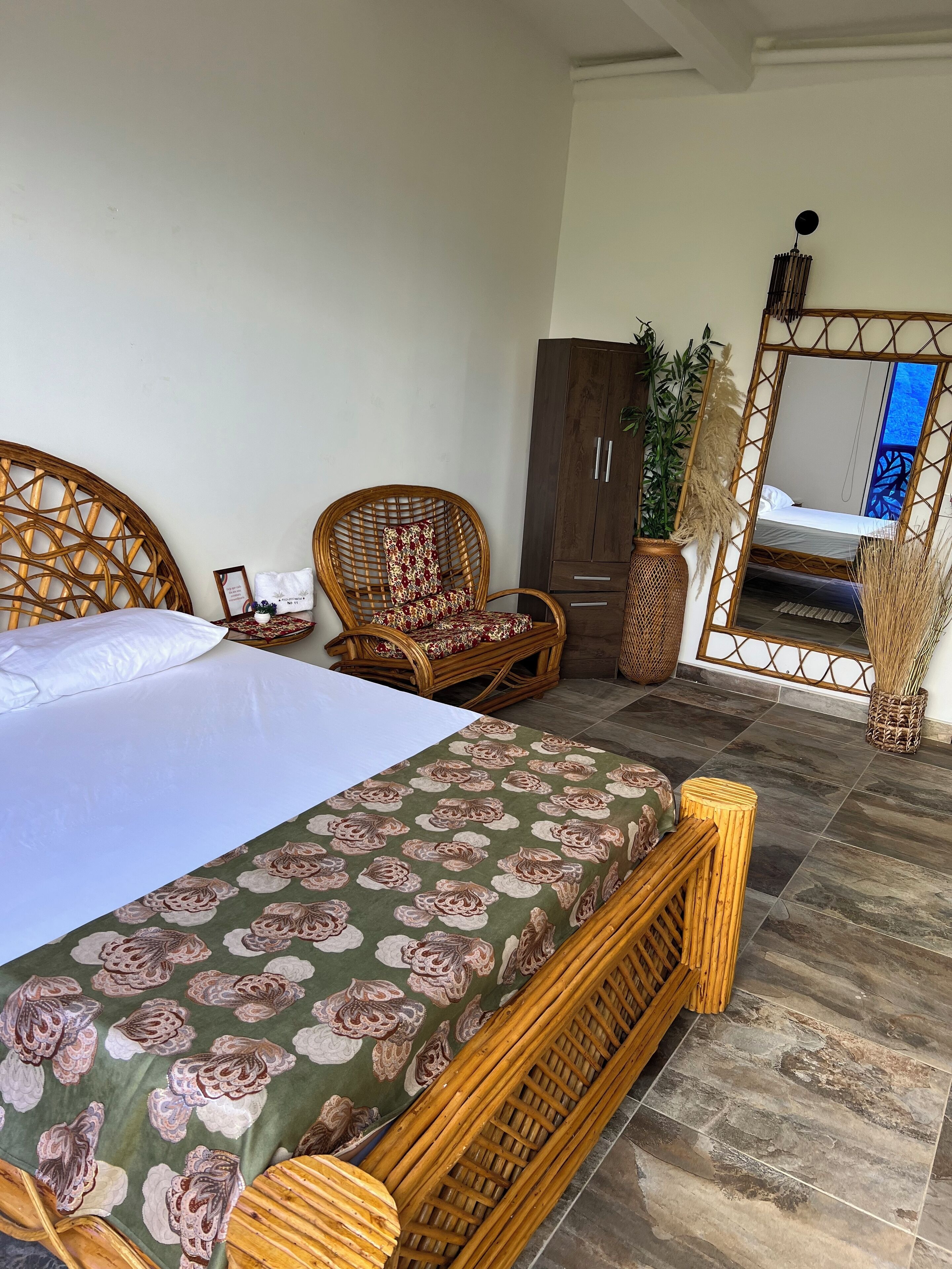 Un dormitorio con cama, una silla de mimbre, un armario y un espejo que refleja una cama.