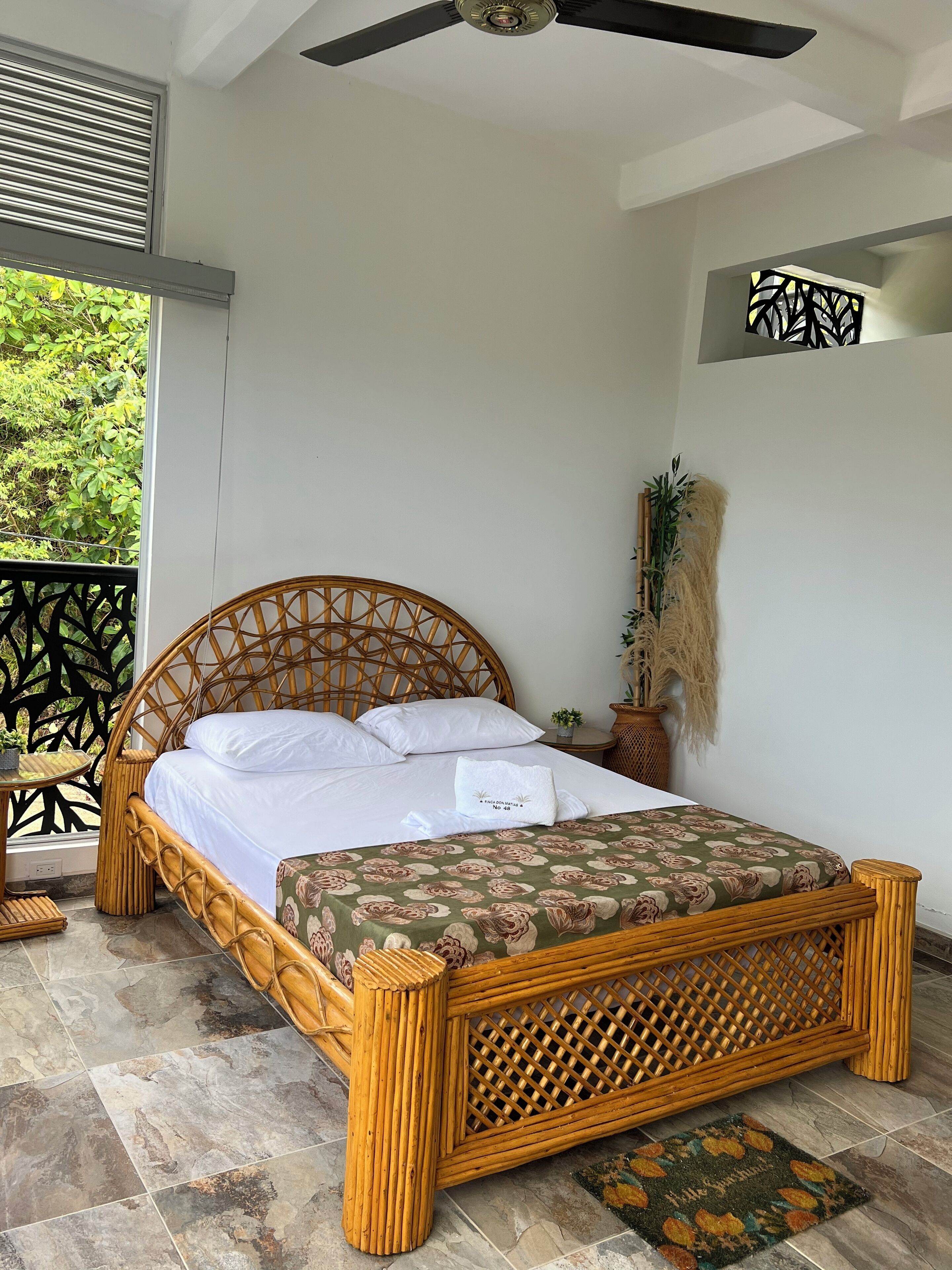 A room with a bed, a ceiling fan, a window with blinds, a potted plant, and a decorative wall piece.