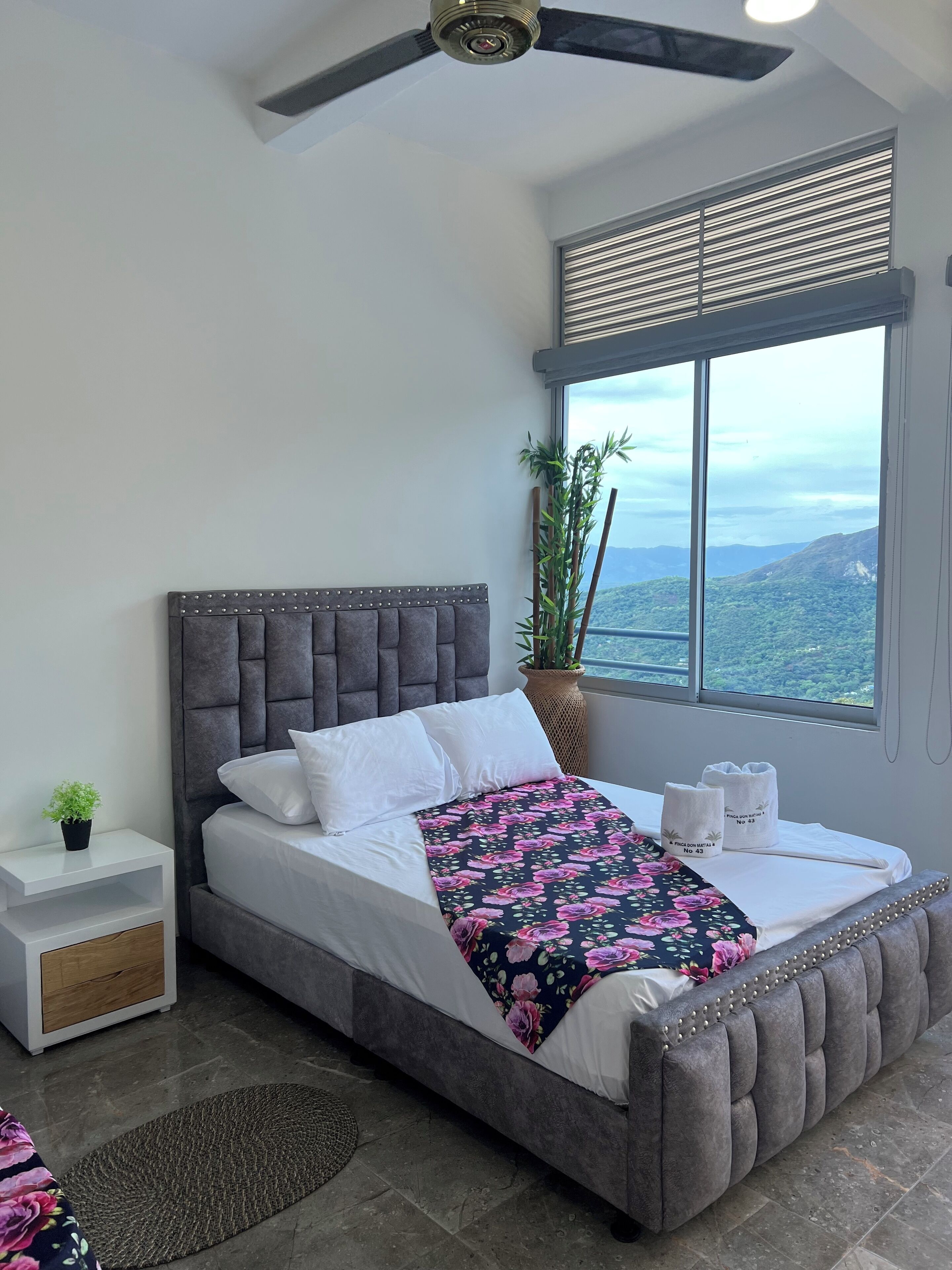Un dormitorio con una cama grande, un ventilador de techo, una ventana con persianas, una mesita de noche y vistas al exterior.