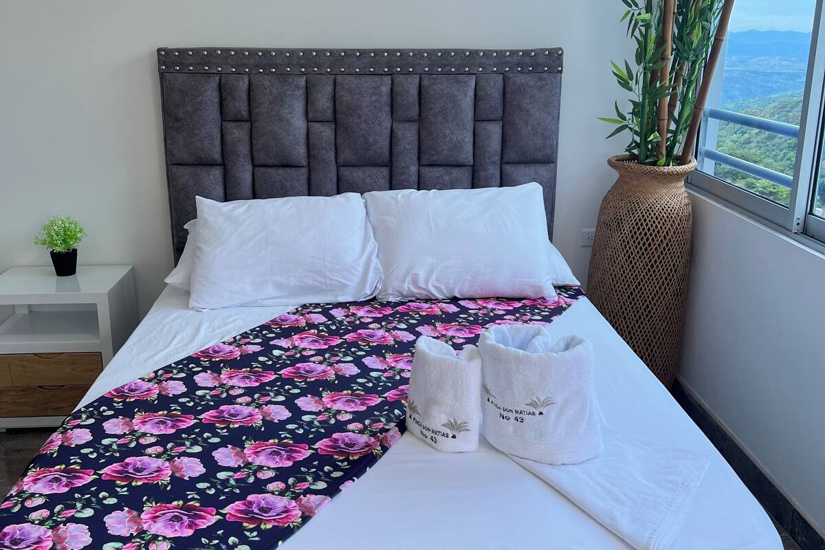 A neatly made bed with a floral bedspread, a grey tufted headboard, and a view of greenery through a window.