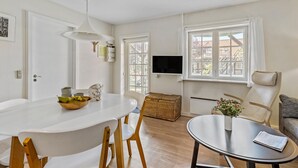 Een moderne woonkamer met een witte eettafel, houten stoelen, een ronde salontafel, een aan de muur bevestigde televisie en grote ramen met uitzicht op gebouwen.