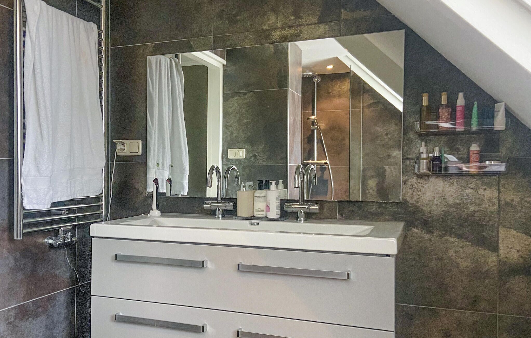 A modern bathroom with a large mirror, a white sink, and a shower. There are toiletries on a shelf and a towel hanging on a rack.
