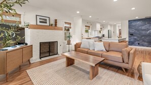 A modern living room with a fireplace, a sofa, a coffee table, and a dining area in the background. - Luxury 3BD 2bath Near EXPO & TU (Tulsa)