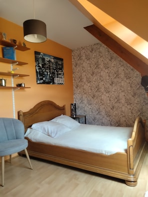A bedroom with a wooden bed, a chair, a shelf with items, a cityscape poster, and a patterned wallpaper.