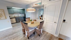 Un coin repas avec une table ronde et des chaises en osier, une cuisine en arrière-plan et une porte blanche avec une serrure.