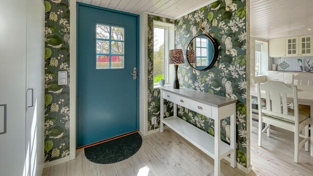 Ein Zimmer mit einer türkisblauen Tür, floralem Wandpapier, einem weißen Konsolentisch, einem runden Spiegel und einem weißen Essbereich mit Holztisch und Stühlen.