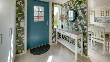 Een kamer met een blauwgroene deur, bloemenbehang, een witte console tafel, een ronde spiegel en een wit eetgedeelte met een houten tafel en stoelen.