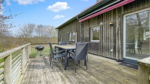 Eine Holzdiele mit Tisch, Stühlen, einem Grill und einem Fenster mit Blick auf eine natürliche Landschaft.