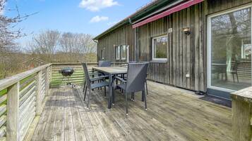 Een houten terras met een tafel, stoelen, een barbecue en een raam met uitzicht op een natuurlandschap.