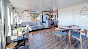 A modern living room with a grey sofa, wooden coffee table, and blue chairs. A kitchen with a fridge and microwave is visible in the background. - Beachcomber: Beachfront Escape Unit #4 (Florence)