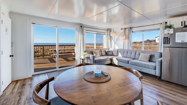 A spacious living area with a large wooden table, a grey sofa, and a view of the beach through large sliding doors. - Beachcomber: Beachfront Escape Unit #4 (Florence)