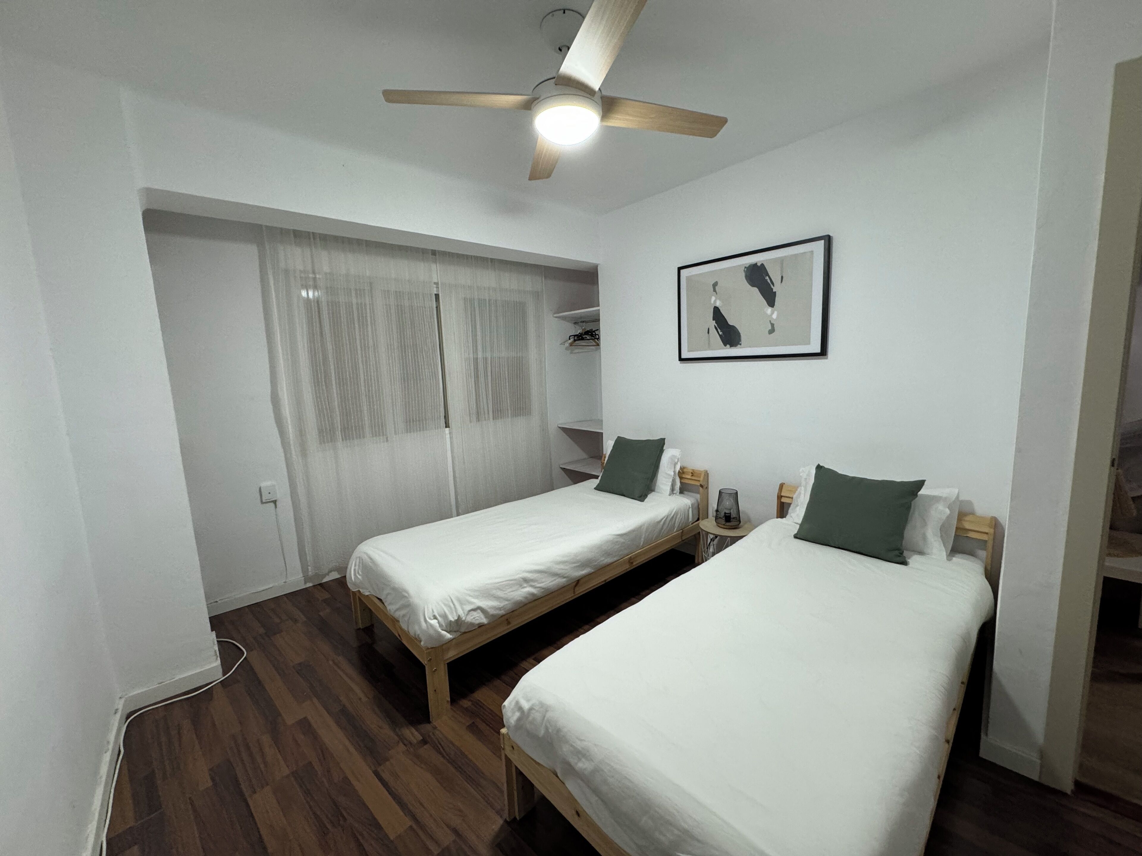Una habitación con dos camas individuales, un ventilador de techo, un cuadro enmarcado en la pared y piso de madera.
