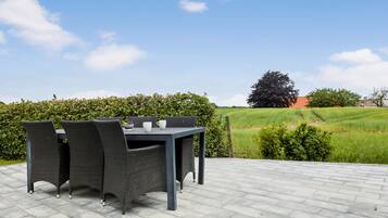 An outdoor patio with a table and chairs, a bowl of fruit, and a view of a green field and trees.