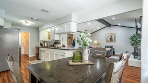 A modern kitchen with white cabinets, a wooden dining table, and a dining area with chairs. - Country Pool House w/ Game Room, Fire Pit, Grill (North Richland Hills)