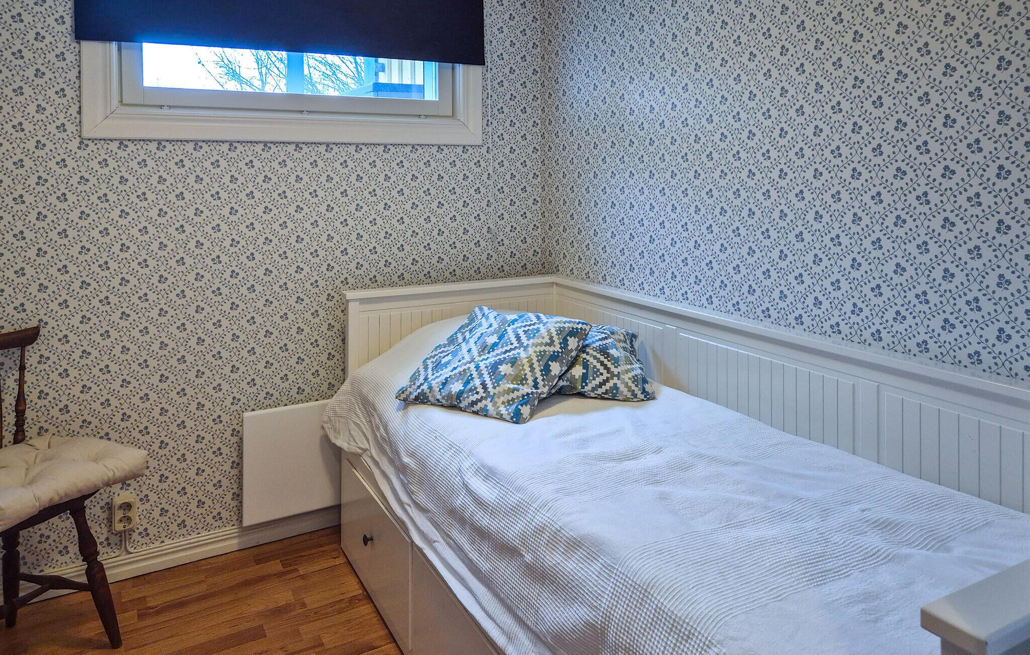 A small bedroom with a single bed, a window with a black blind, and a wooden chair.