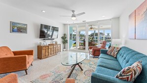 A modern living room with a glass coffee table, orange and blue sofas, a wooden cabinet, and a TV. - Canal-Front Home with Heated Pool (Cocoa Beach)