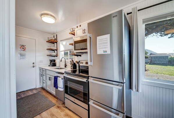 A compact kitchen with a stainless steel refrigerator, oven, and built-in microwave. There are wooden shelves above the counter, and a window with a view of the outdoors. - Beachcomber: Beachfront Escape Unit #5 (Florence)