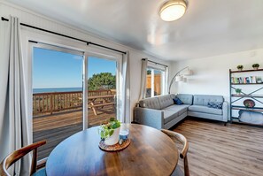 A modern living room with a wooden floor, a grey sofa, a round table with a plant, and a balcony with a wooden deck and railing. - Beachcomber: Beachfront Escape Unit #5 (Florence)