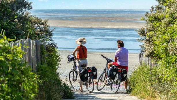 Deux personnes à vélo se trouvent à l’entrée d’une plage, de part et d’autre d’une clôture en bois et d’une végétation luxuriante.