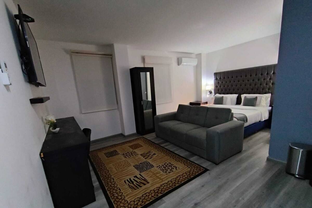 Una habitación de hotel moderna con un sofá gris, una cama con cabecera capitonada, una mesita y una alfombra decorativa.