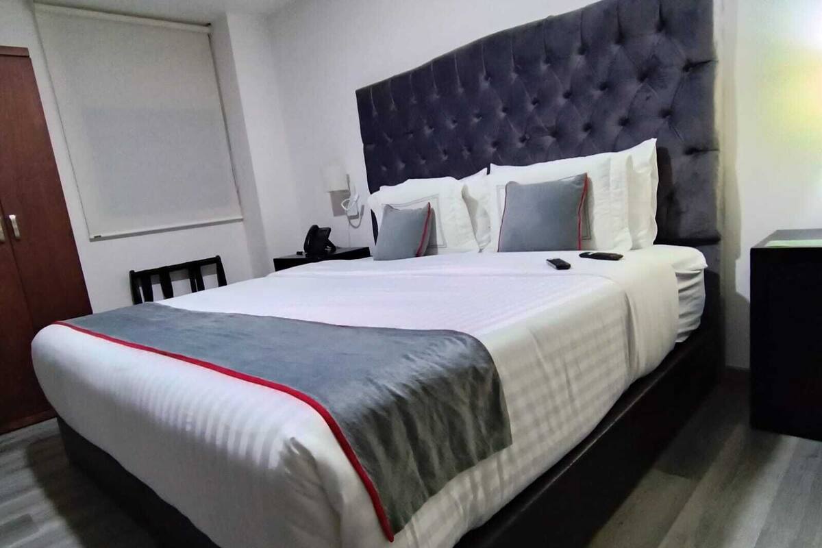 Una habitación de hotel con una cama grande, un cabecero oscuro y una mesita de noche con una lámpara.
