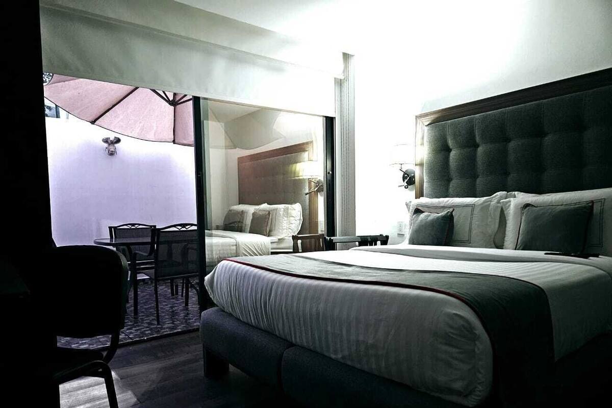 Habitación de hotel con una cama grande, un escritorio y una silla. Posee una puerta de vidrio corrediza que da a un espacio exterior con una mesa y sillas.
