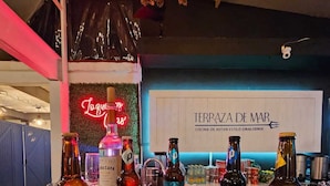 Zona de bar con diversas botellas de licores, incluyendo IPA y picudo, y un letrero de neón que dice "Terraza de Mar".