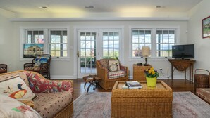 A living room with a fireplace, wicker furniture, a television, and a large window with an American flag. - Duplex in Lexington, VA, Loranwoods (Rockbridge County)
