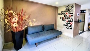 Un moderno vestíbulo de hotel con un sofá gris, una planta decorativa y una pared con saludos en varios idiomas.