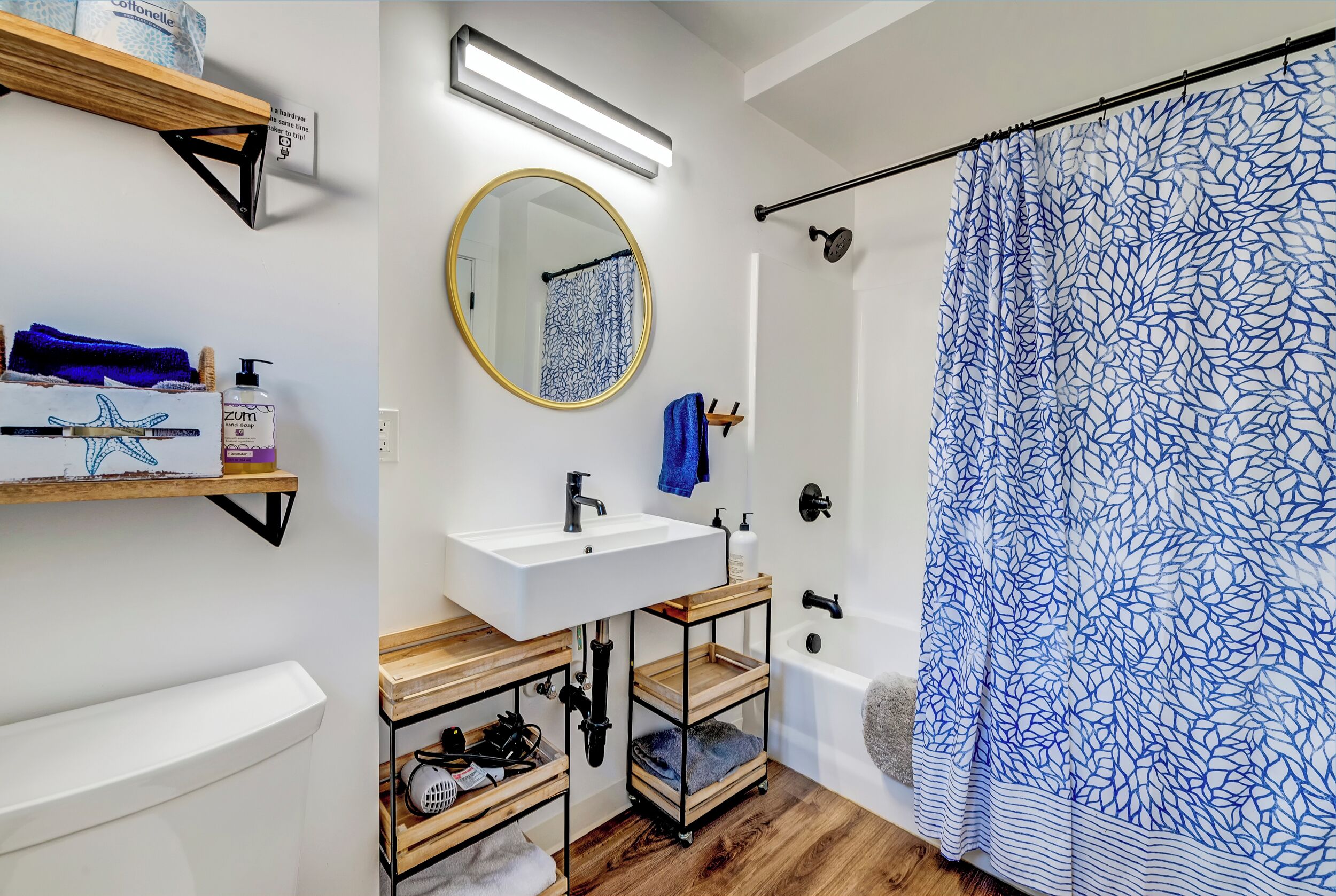 A bathroom with a white sink, wooden shelves, a round mirror, and a shower with a blue patterned curtain.
