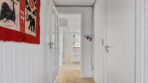 Un couloir étroit aux murs blancs et au plancher en bois, qui mène à un espace cuisine doté d’une fenêtre et d’une lampe suspendue.