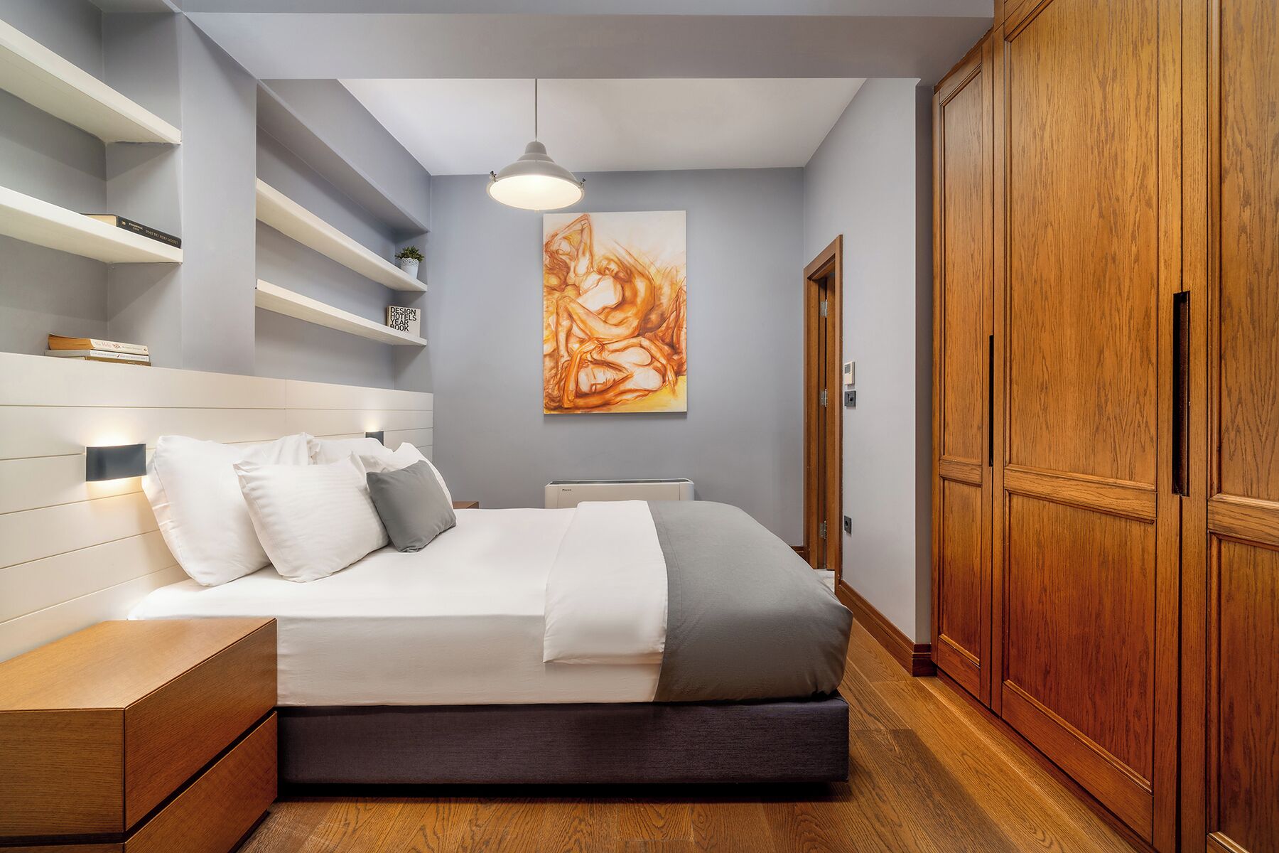 A bedroom with a bed, bedside tables, a wardrobe, and a wall-mounted shelf with books and a plant.