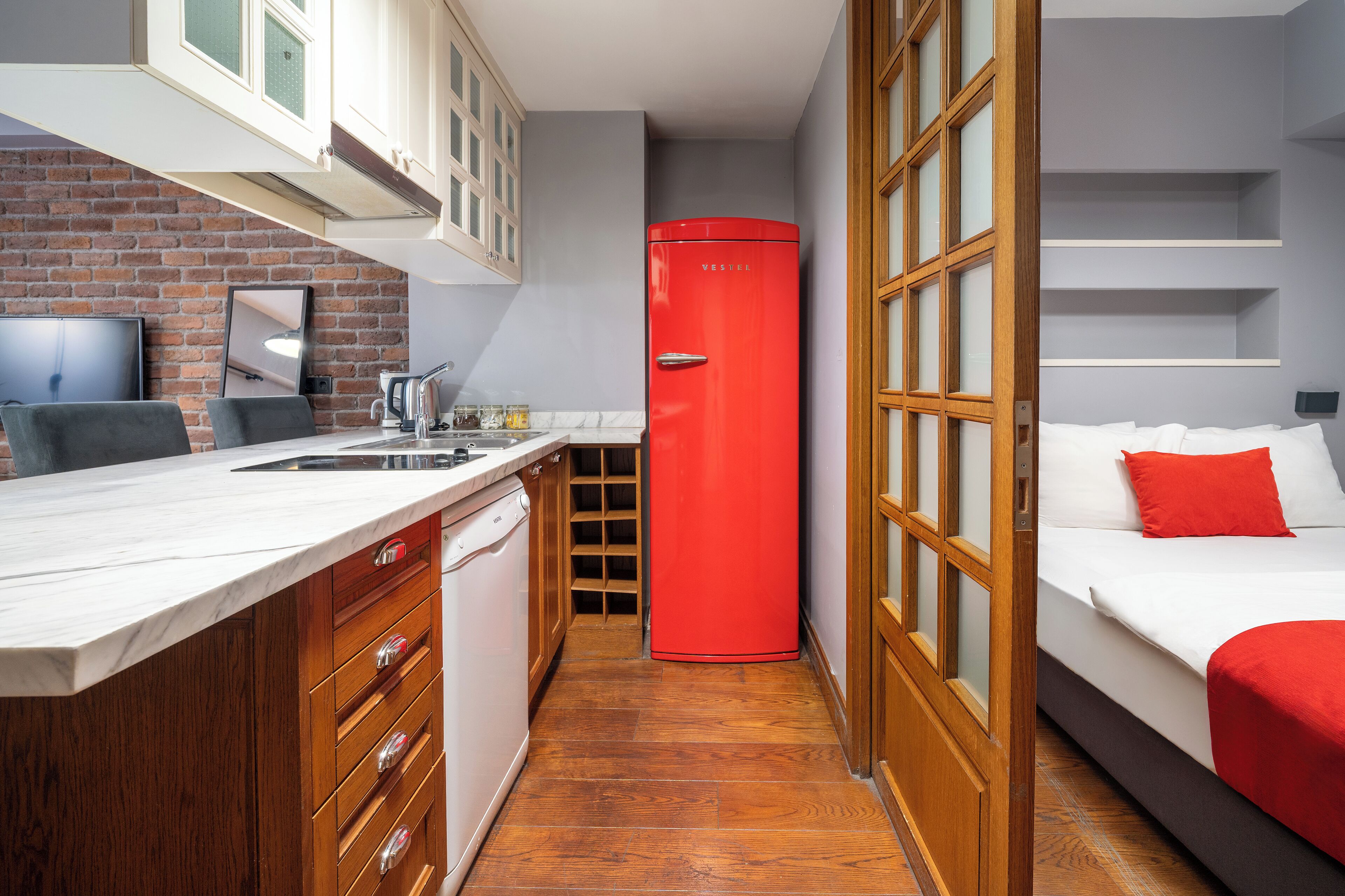 A compact hotel room with a kitchen area, a red refrigerator, a bed with red bedding, and wooden cabinets.