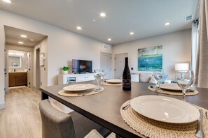 Un comedor moderno con una larga mesa dispuesta para cuatro personas, una botella de vino y un televisor al fondo.
