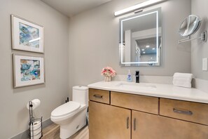 Un baño moderno con un lavamanos blanco, un espejo con luz, una obra de arte enmarcada en la pared y un inodoro.