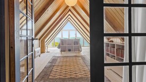 Un dormitorio en el ático con vigas de madera, un ventanal grande, una estantería y una lámpara colgante.