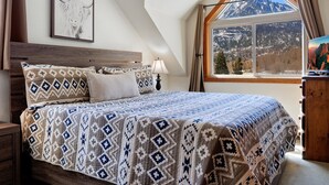Une chambre dotée d’un grand lit, d’une tête de lit en bois, d’une fenêtre donnant sur les montagnes et d’une œuvre d’art encadrée accrochée au mur.