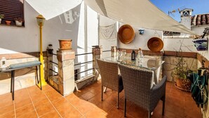 Een terras buiten met een tafel, stoelen, een gele lamp en potplanten.
